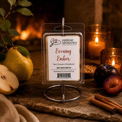 A package of Jarhead Aromatics Evening Ember Soy Wax Melt | Plum, Sandalwood & Soft Smoke is displayed on a stand, surrounded by pears, plums, cinnamon sticks, a lit candle, and warm ambient lighting. The inviting arrangement perfectly captures the comforting essence of these premium soy wax melts from Jarhead Aromatics.