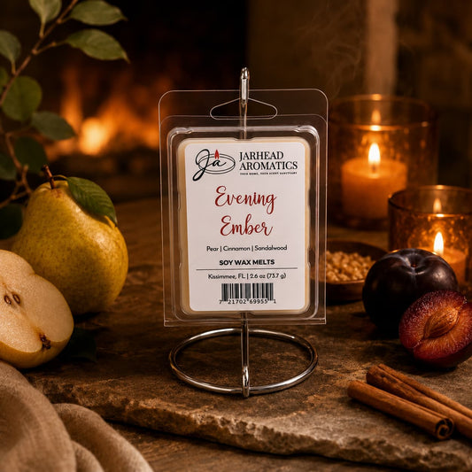 A package of Jarhead Aromatics Evening Ember Soy Wax Melt | Plum, Sandalwood & Soft Smoke is displayed on a stand, surrounded by pears, plums, cinnamon sticks, a lit candle, and warm ambient lighting. The inviting arrangement perfectly captures the comforting essence of these premium soy wax melts from Jarhead Aromatics.