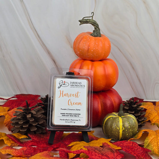 A stack of three pumpkins rests on autumn leaves with pinecones and a small green pumpkin nearby. In front, a package of Harvest Cream - Soy Wax Melt by Jarhead Aromatics is displayed on a small black easel.