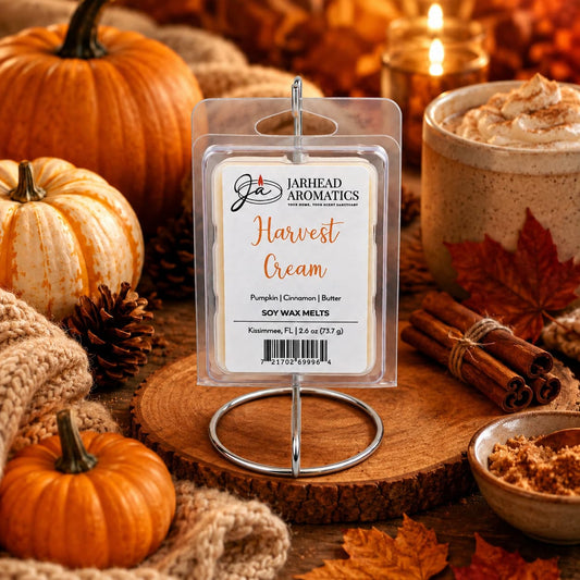A package of Jarhead Aromatics Harvest Cream Soy Wax Melt | Pumpkin Spice, Brown Sugar & Vanilla Cream is displayed on a stand, surrounded by pumpkins, a candle, a mug of whipped drink, cinnamon sticks, and autumn leaves in a cozy fall setting. Fill your home with the inviting aroma and long-lasting fragrance of pumpkin spice, brown sugar, and vanilla cream from Jarhead Aromatics.