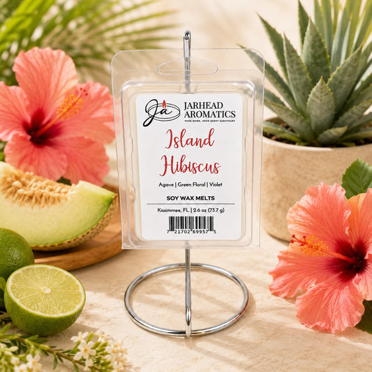A package of Jarhead Aromatics Island Hibiscus Soy Wax Melt | Honeydew, Lime & Tropical Florals rests on a metal holder, surrounded by vibrant hibiscus blooms, a halved lime, and potted succulents on a light surface—infusing your space with the essence of tropical home fragrance.