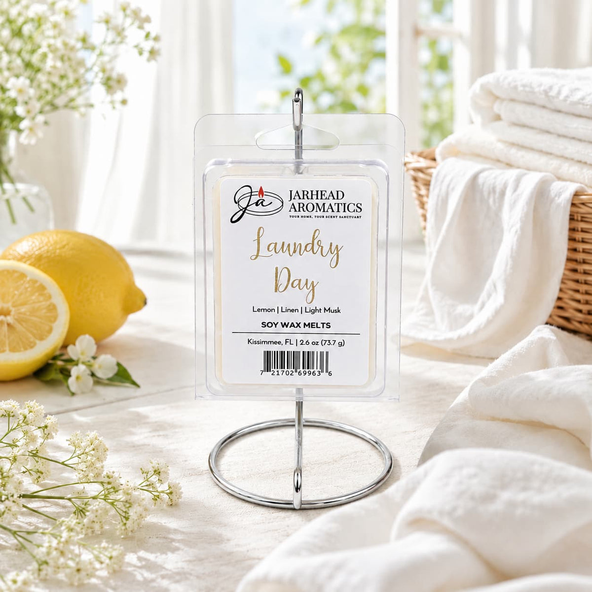 A package of Jarhead Aromatics Laundry Day Soy Wax Melt | Fresh Linen, Lemon & Clean Musk is displayed on a metal stand, surrounded by fresh lemons, white towels, and sunlight in the background. The scene evokes a crisp and clean atmosphere, perfectly capturing the essence of this home fragrance that infuses any space with notes of fresh linen, zesty lemon, and clean musk.