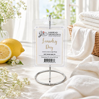 A package of Jarhead Aromatics Laundry Day Soy Wax Melt | Fresh Linen, Lemon & Clean Musk is displayed on a metal stand, surrounded by fresh lemons, white towels, and sunlight in the background. The scene evokes a crisp and clean atmosphere, perfectly capturing the essence of this home fragrance that infuses any space with notes of fresh linen, zesty lemon, and clean musk.