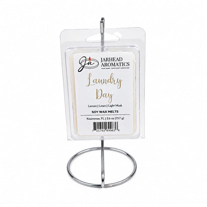 A package of Jarhead Aromatics’ Laundry Day Soy Wax Melt | Fresh Linen, Lemon & Clean Musk rests gracefully on a metal stand. The transparent packaging highlights a sophisticated white and gold label featuring the scent notes: Fresh Linen, Lemon, and Clean Musk. Ideal for anyone who loves home fragrance, these soy wax melts envelop your space in the revitalizing aroma of freshly laundered linens mixed with bright lemon and clean musk—bringing the crisp essence of laundry day into any room.