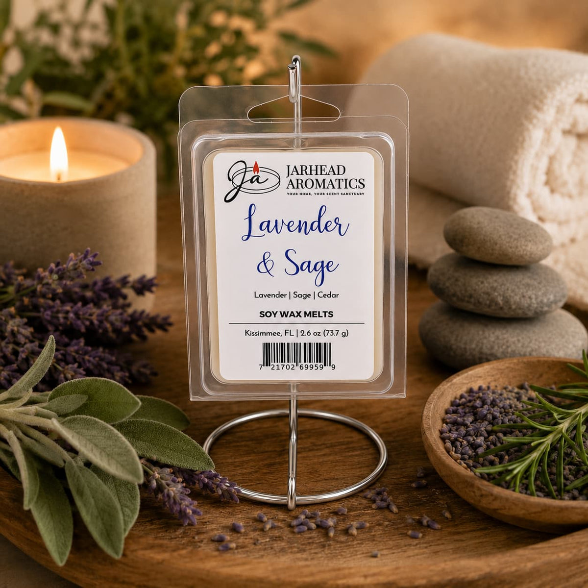 A pack of Jarhead Aromatics Lavender & Sage Soy Wax Melt | Lavender, Sage & Sandalwood is displayed alongside lavender sprigs, smooth stones, a lit candle, and a rolled towel, evoking a soothing spa-like atmosphere. This herbal soy wax melt blend fills your home with the calming aroma of lavender, sage, and sandalwood for true tranquility.