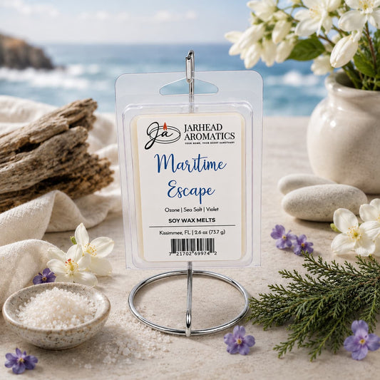 A package of Jarhead Aromatics Maritime Escape Soy Wax Melt | Sea Salt, Ozone & Jasmine is displayed on a metal stand, surrounded by white flowers, salt, and sand with a coastal ocean view in the background—inviting you to enjoy the refreshing fragrance of ocean air blended with sea salt, ozone, jasmine, and hints of coastal woods.