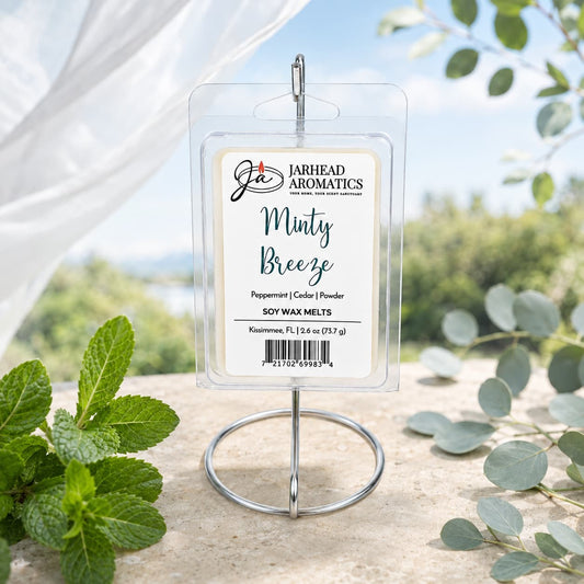 A package of Jarhead Aromatics Minty Breeze Soy Wax Melt | Peppermint, Eucalyptus & Cedarwood rests on a stand outdoors, encircled by fresh mint leaves and eucalyptus with a softly blurred natural backdrop. Infused with an invigorating blend of peppermint, eucalyptus, and cedarwood fragrance, these soy wax melts from Jarhead Aromatics fill your home with a soothing scent ideal for any room.