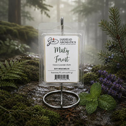 A package of Jarhead Aromatics Misty Forest Soy Wax Melt | Moss, Juniper & Rain-Kissed Woods is showcased on a metal stand in a misty forest, surrounded by green moss, ferns, and purple flowers, capturing the essence of natural home fragrance with its enchanting blend of moss, juniper, and rain-kissed woods.