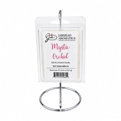 A package of Jarhead Aromatics Mystic Orchid Soy Wax Melt | Orchid, Salt Air & Vanilla is displayed on a metal stand. The label highlights scents of orchid, salt air, and vanilla for a true coastal home fragrance experience. The packaging features white and pink text and showcases the premium soy wax inside through its clear wrapping.