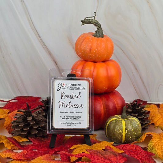 A display of pumpkins, pinecones, and autumn leaves surrounds the Jarhead Aromatics Roasted Molasses - Soy Wax Melt from the Fall Collection, filling the air with a rich caramelized molasses aroma atop a small black easel.
