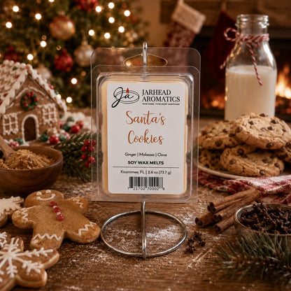 A Jarhead Aromatics Santa’s Cookies Soy Wax Melt package is displayed on a metal stand, surrounded by gingerbread cookies, chocolate chip cookies, and a bottle of milk to evoke a holiday baking fragrance.