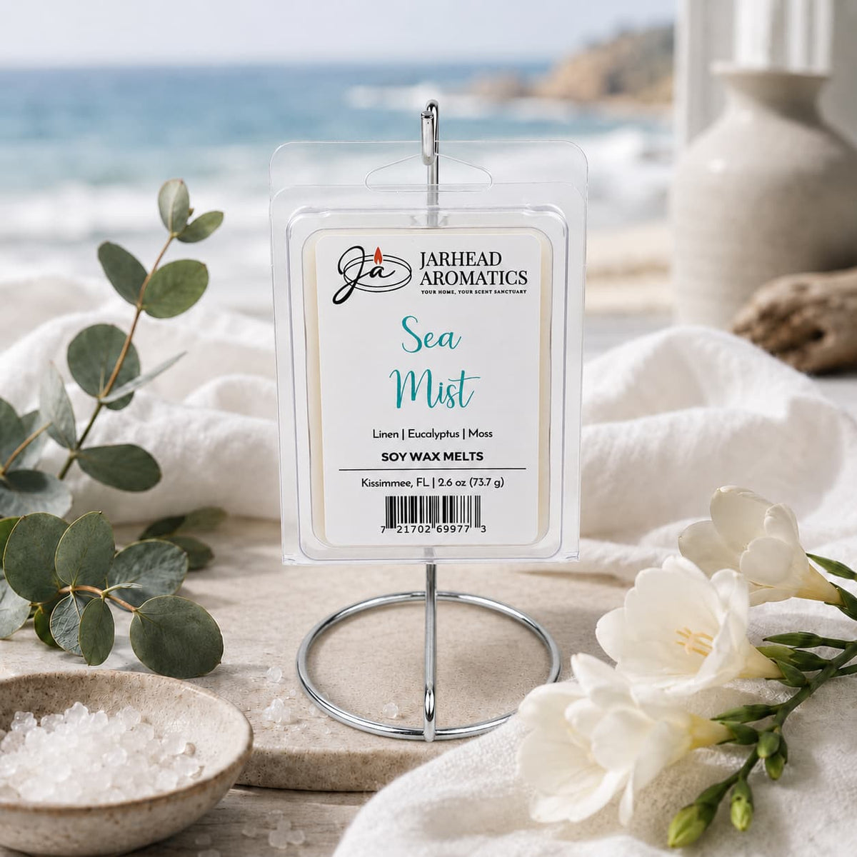 The Jarhead Aromatics Sea Mist Soy Wax Melt | Fresh Linen, Sea Salt & Eucalyptus is displayed on a stand, surrounded by white flowers, eucalyptus, and sea salt, with a beach and ocean visible through a window in the background. This spa-inspired melt fills your home with the refreshing and serene fragrance of fresh linen, sea salt, and eucalyptus—evoking the tranquility of the shoreline.