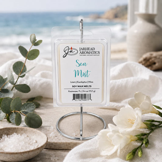 The Jarhead Aromatics Sea Mist Soy Wax Melt | Fresh Linen, Sea Salt & Eucalyptus is displayed on a stand, surrounded by white flowers, eucalyptus, and sea salt, with a beach and ocean visible through a window in the background. This spa-inspired melt fills your home with the refreshing and serene fragrance of fresh linen, sea salt, and eucalyptus—evoking the tranquility of the shoreline.