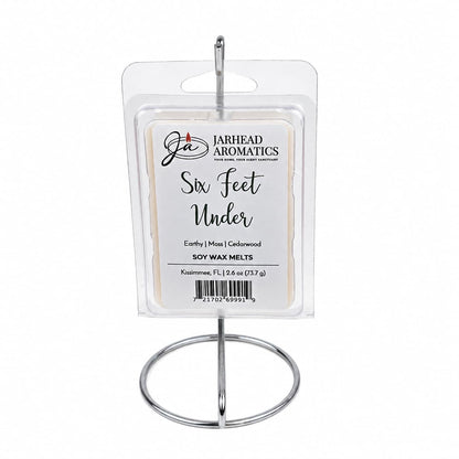 A package of Jarhead Aromatics’ Six Feet Under Soy Wax Melt | Damp Earth, Moss & Ancient Oak is displayed on a silver stand. The clear, rectangular packaging features a label that highlights its damp earth, moss, and ancient oak scents. This dark and atmospheric fragrance truly captures the essence of Six Feet Under, filling any space with a mysterious and captivating aroma.