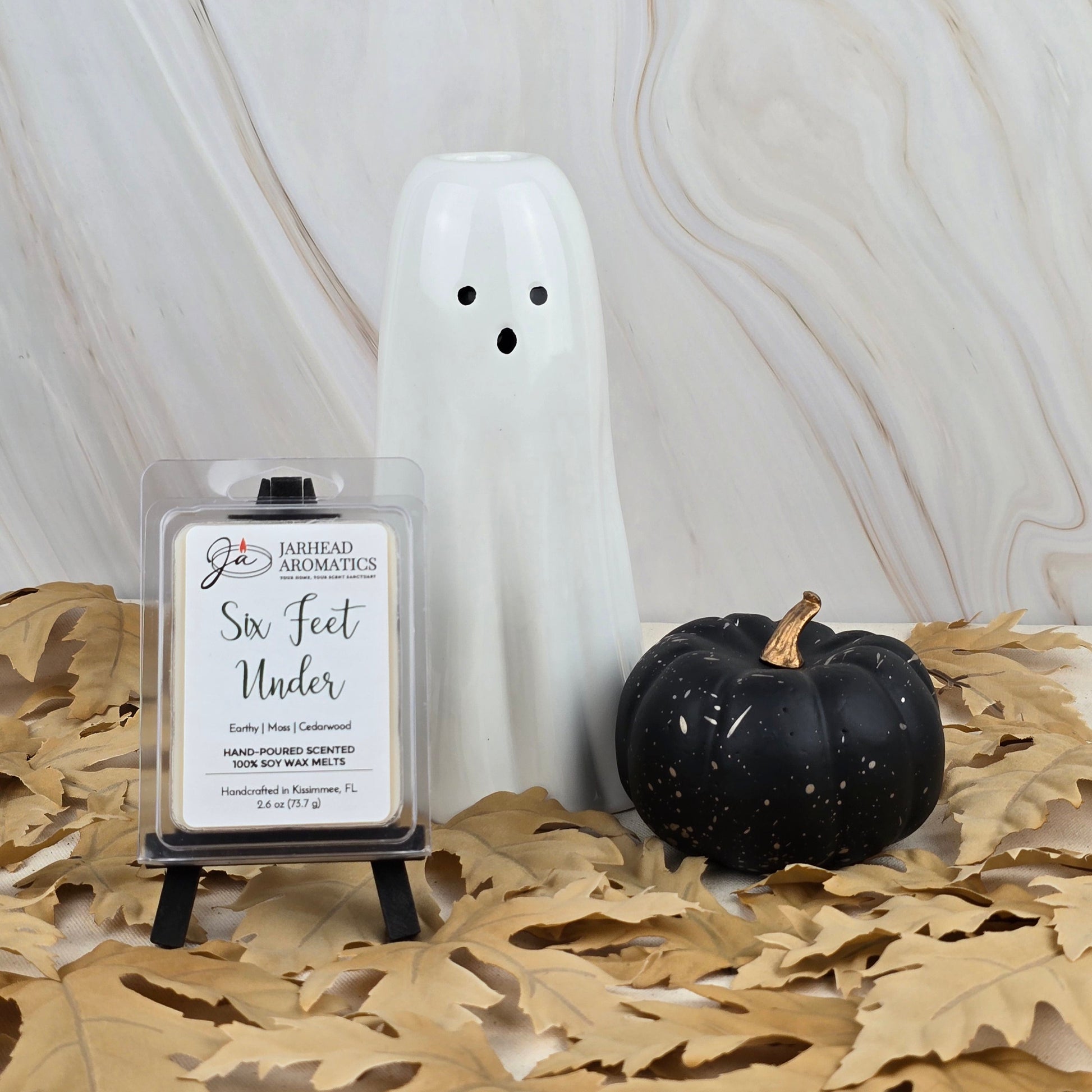 A white ghost figurine, a black pumpkin, and Jarhead Aromatics’ Six Feet Under Soy Wax Melt from the Halloween Collection with an old cemetery scent rest on dried leaves against a marble background.