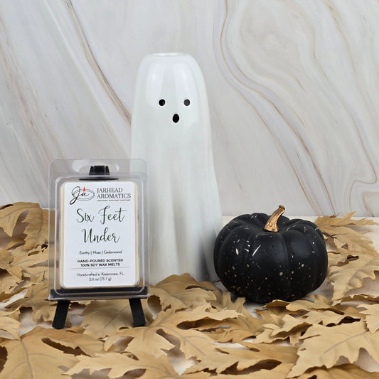 A white ghost figurine, a black pumpkin, and Jarhead Aromatics’ Six Feet Under Soy Wax Melt from the Halloween Collection with an old cemetery scent rest on dried leaves against a marble background.