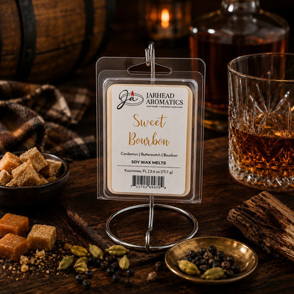 A package of Jarhead Aromatics’ Sweet Bourbon Soy Wax Melt | Bourbon, Butterscotch & Brown Sugar—a gourmand blend celebrated for its long-lasting scent—is showcased on a metal stand, surrounded by a glass of bourbon, brown sugar cubes, cardamom pods, peppercorns, and butterscotch atop a rustic wooden table.