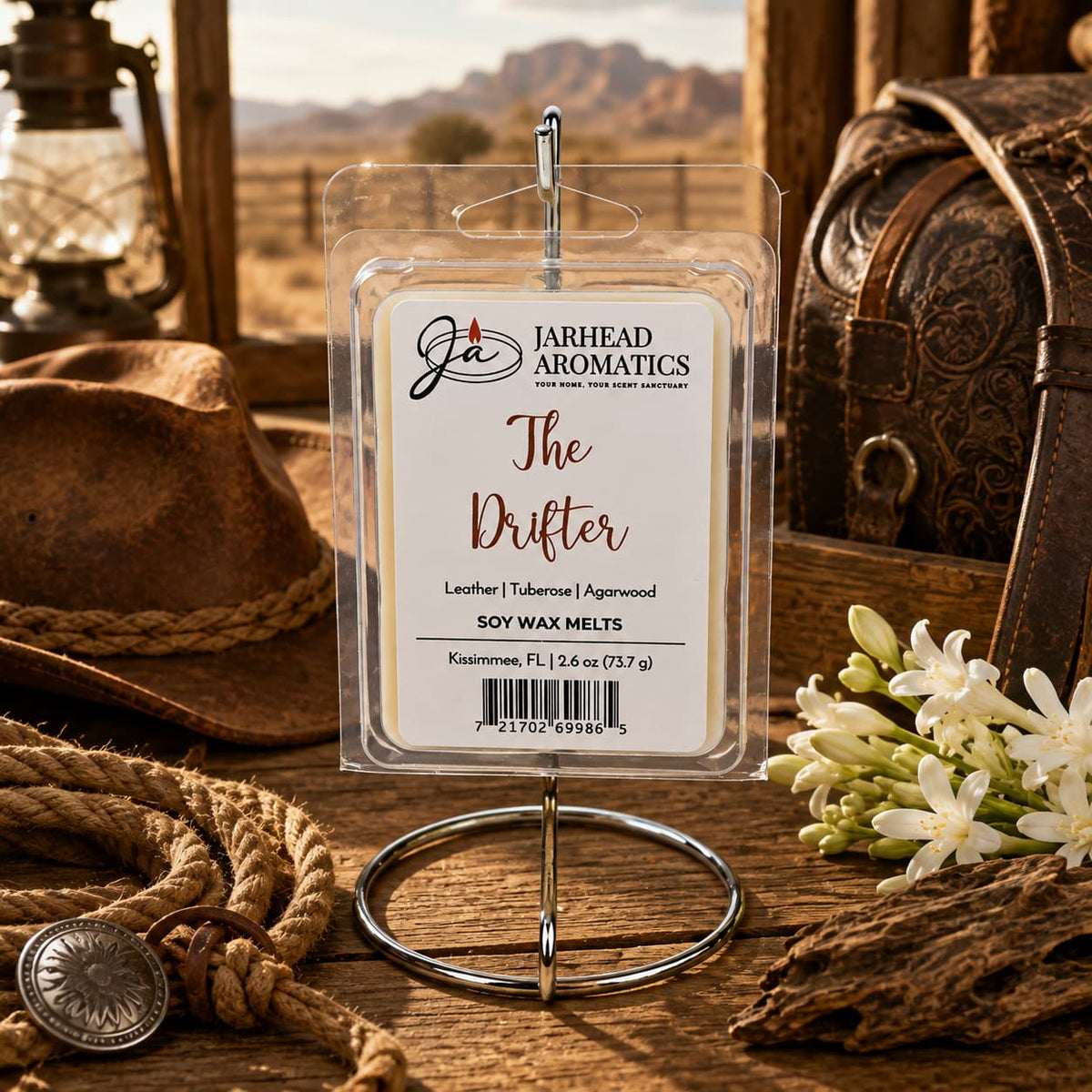 A pack of Jarhead Aromatics The Drifter Soy Wax Melt | Leather, Agarwood & Tuberose rests on a metal stand amidst cowboy gear, coiled rope, a lantern, white flowers, and a leather bag in a rustic, Old West-inspired setting. The scene captures the rugged allure of frontier days while hinting at the rich blend of leather, agarwood, and tuberose that infuses each handcrafted soy wax melt.