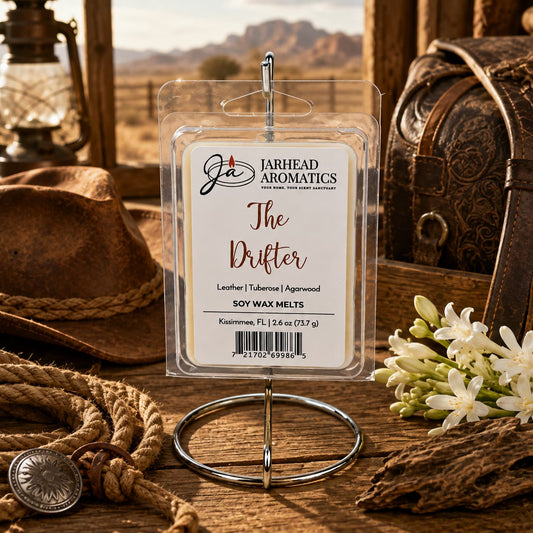 A pack of Jarhead Aromatics The Drifter Soy Wax Melt | Leather, Agarwood & Tuberose rests on a metal stand amidst cowboy gear, coiled rope, a lantern, white flowers, and a leather bag in a rustic, Old West-inspired setting. The scene captures the rugged allure of frontier days while hinting at the rich blend of leather, agarwood, and tuberose that infuses each handcrafted soy wax melt.