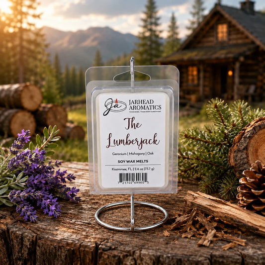 A package of The Lumberjack Soy Wax Melt by Jarhead Aromatics is displayed on a holder outdoors, surrounded by lavender, pinecones, logs, and a log cabin against a backdrop of trees and mountains at sunset. The scene perfectly captures the woodsy fragrance blend of mahogany, cedarwood, and oak that these soy wax melts evoke.