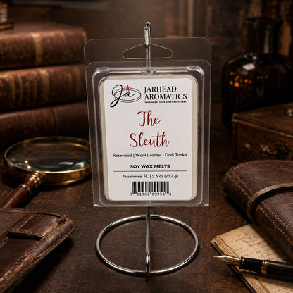A package of The Sleuth Soy Wax Melt | Leather, Rosewood & Sandalwood by Jarhead Aromatics is showcased on a stand amid vintage books, a quill, and antique decor atop a wooden desk—ideal for infusing your home fragrance collection with an air of mystery and sophistication.