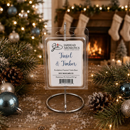 A package of Tinsel & Timber Soy Wax Melt | Fresh Pine Christmas Tree Holiday Scent in a circular wire stand sits on a wooden surface decorated with pine branches, pinecones, and Christmas ornaments in front of a fireplace.
