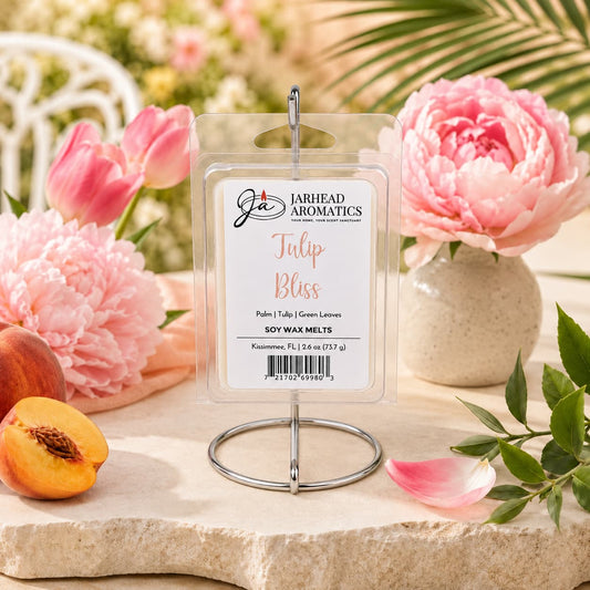 A package of Jarhead Aromatics Tulip Bliss Soy Wax Melt | Tulip, Peony & Sweet Peach is displayed on a stand, encircled by vibrant pink peonies, lush green leaves, and a halved peach, evoking a fresh and summery atmosphere with its inviting floral home fragrance.
