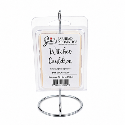 A package of Jarhead Aromatics Witches Cauldron Soy Wax Melt | Orange Blossom, Patchouli & Dark Incense sits atop a silver display stand. The label showcases notes of orange blossom, patchouli, and dark incense for a highly fragrant experience, and features a barcode at the bottom.