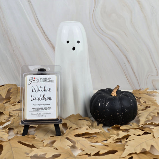 A white ceramic ghost, a small black pumpkin, and Jarhead Aromatics Witches Cauldron Soy Wax Melt from the Halloween Collection sit on brown leaves against a marbled background.