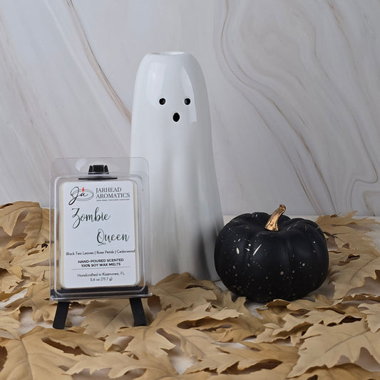 A white ceramic ghost, a black pumpkin, and a pack of “Zombie Queen - Soy Wax Melt” by Jarhead Aromatics are arranged on tan leaves atop marble.