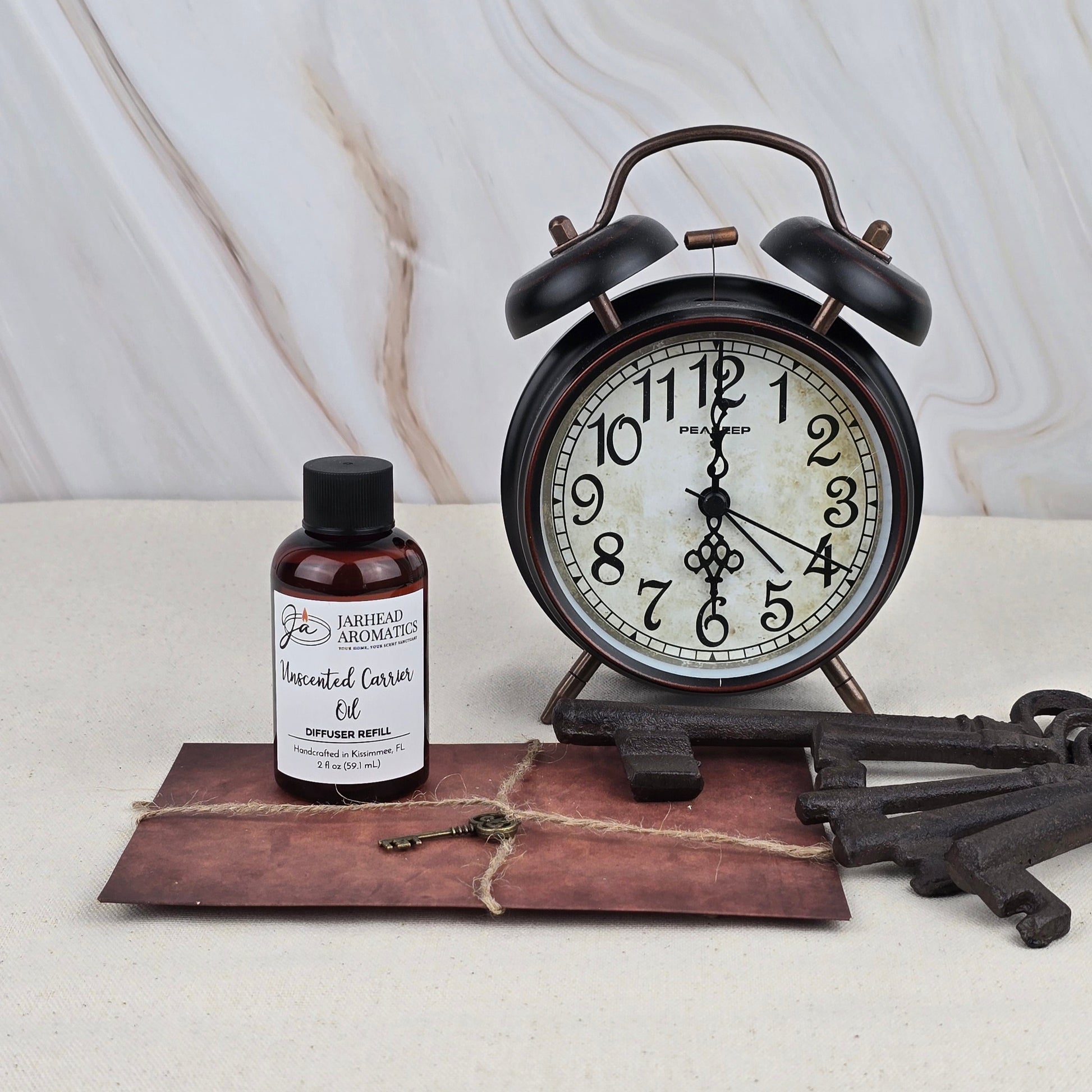 A black vintage alarm clock set to 9:55 sits beside a Jarhead Aromatics Unscented Carrier Oil - Diffuser Refill, a brown string-tied envelope, and three large antique keys, all displayed on a light surface with a marble backdrop.