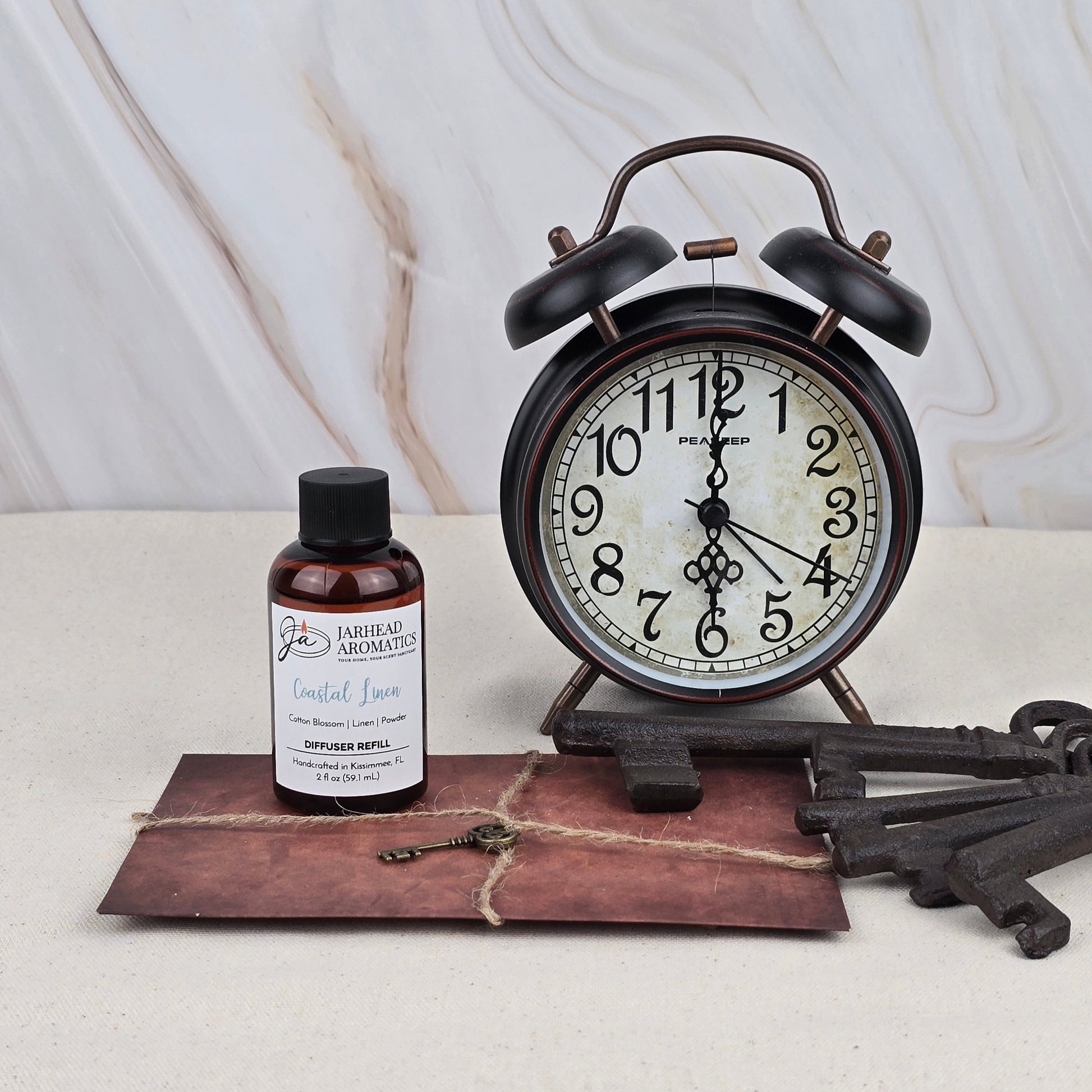 A vintage black alarm clock shows 10:09 on a table with Jarhead Aromatics’ Coastal Linen Diffuser Refill, two antique keys, and a sealed brown envelope tied with string, set against a light marble background.