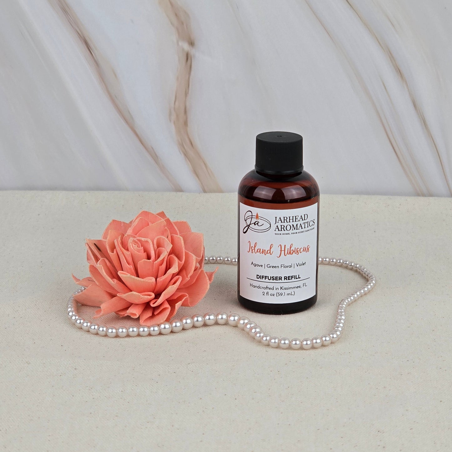 A bottle of Jarhead Aromatics Island Hibiscus - Diffuser Refill rests on a light surface beside a peach artificial flower and pearls, infusing the air with tropical fragrance against a marble-patterned backdrop.