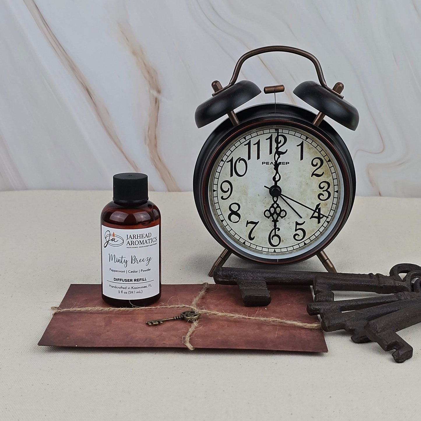 A vintage black alarm clock, a large metal key, a tied brown envelope, and Jarhead Aromatics’ “Minty Breeze” Diffuser Refill rest on a light surface against a marbled background.