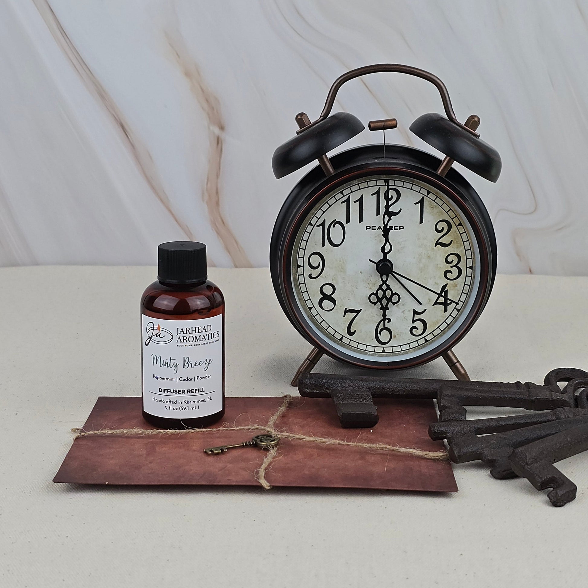 A vintage black alarm clock, a large metal key, a tied brown envelope, and Jarhead Aromatics’ “Minty Breeze” Diffuser Refill rest on a light surface against a marbled background.