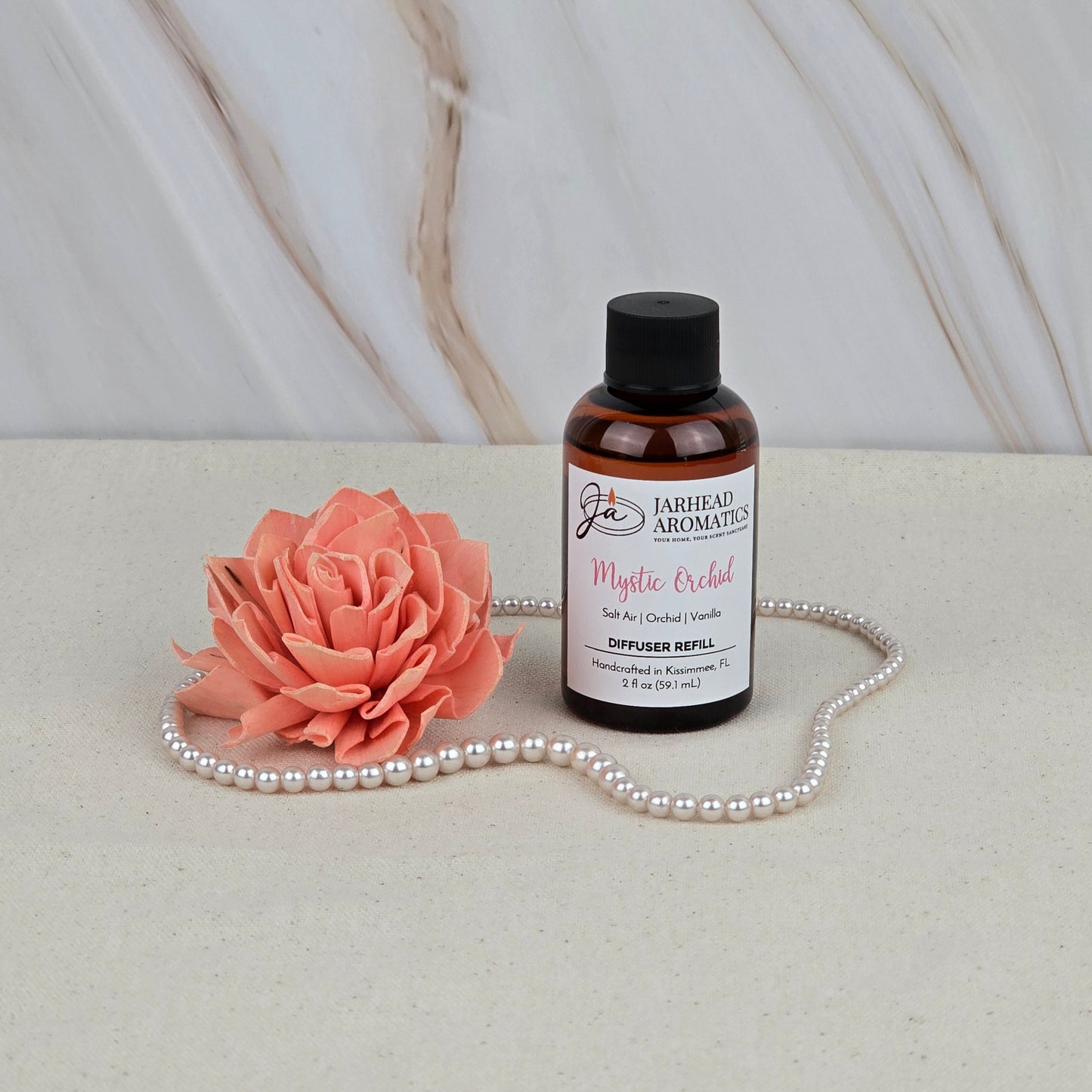 A bottle of Jarhead Aromatics Mystic Orchid - Diffuser Refill sits beside a peach artificial flower and pearls on beige fabric, adding coastal freshness with its fragrance against a marble backdrop.