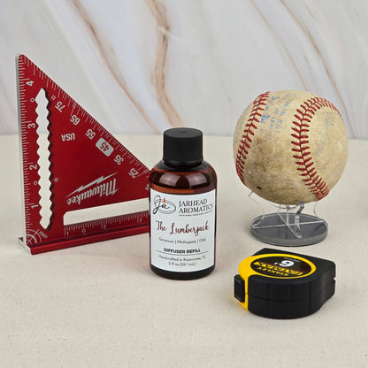 A red triangular ruler, an old baseball, a yellow-black tape measure, and a Jarhead Aromatics The Lumberjack - Diffuser Refill are arranged on beige with a marble backdrop, creating an outdoors-inspired scene.