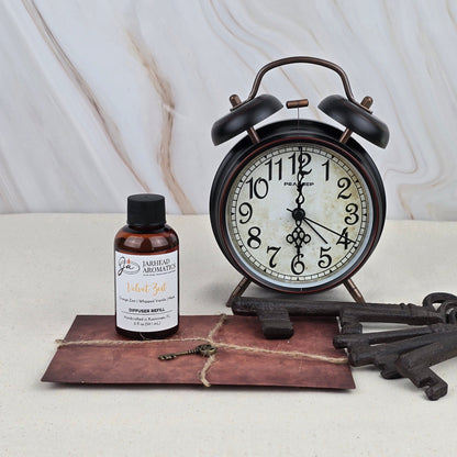 A vintage black alarm clock shows 8:59. Nearby are a Velvet Zest - Diffuser Refill by Jarhead Aromatics in a small brown bottle, a sealed brown envelope with string, and two large antique keys on a light surface with marble background.