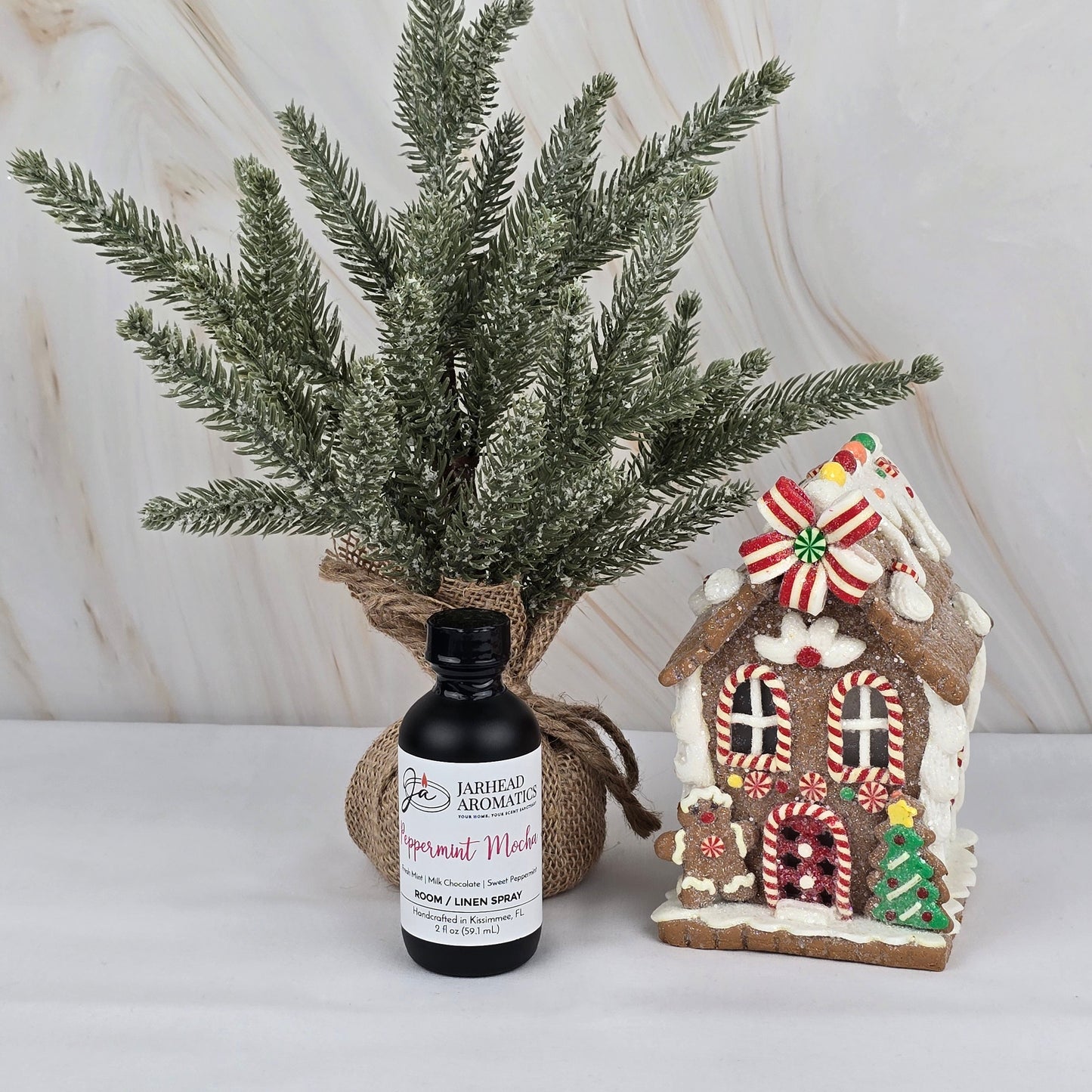 A small frosted pine tree in burlap sits beside Jarhead Aromatics’ Peppermint Mocha Room/Linen Spray from the Winter Collection and a decorated candy-accented gingerbread house, all arranged on a white surface with a marble background.