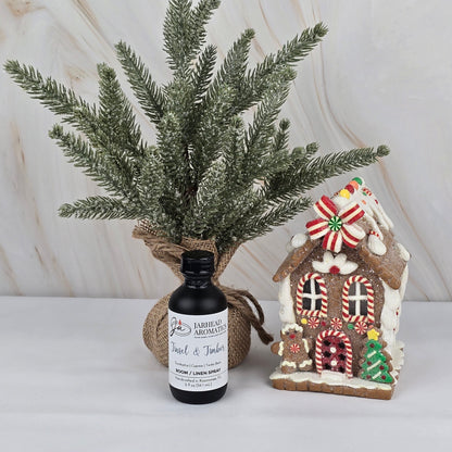 A small frosted pine branch in burlap sits by a gingerbread house decorated with icing and candies. In front, a bottle of Jarhead Aromatics Tinsel & Timber Room / Linen Spray from the Winter Collection adds festive fragrance to the scene.