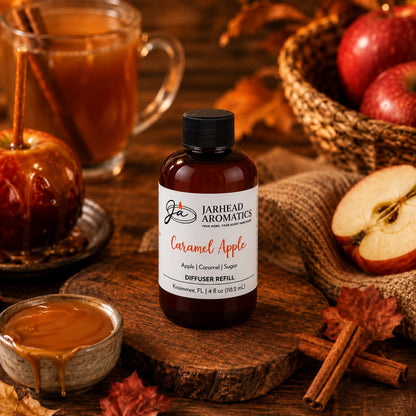 A bottle of Jarhead Aromatics Caramel Apple Diffuser Refill | Crisp Apple, Rich Caramel & Warm Spice rests on a wooden surface alongside caramel, cinnamon sticks, apples, leaves, and a caramel apple, capturing the inviting autumn scent and warm home fragrance of this seasonal blend.