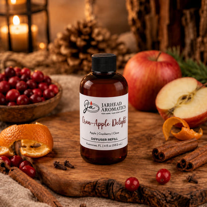 A bottle labeled Cran-Apple Delight Diffuser Refill from Jarhead Aromatics rests on a wooden surface, surrounded by fresh apples, tart cranberries, vibrant orange peels, warm cinnamon sticks, and aromatic cloves—with rustic pinecones completing the inviting scene. In the background, a softly glowing candle enhances the cozy atmosphere, perfectly evoking the comforting blend of cranberry, apple, and warm cinnamon found in this signature home fragrance.
