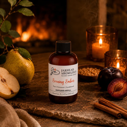 A bottle of Evening Ember Diffuser Refill | Plum, Sandalwood & Soft Smoke from Jarhead Aromatics rests on a table surrounded by pears, plums, cinnamon sticks, and glowing candles. A fireplace radiates warmth in the background, creating a cozy and inviting home fragrance ambiance.