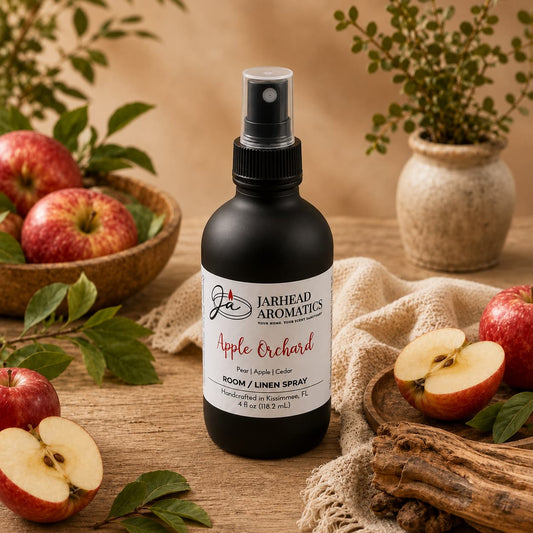 A bottle of Jarhead Aromatics’ Apple Orchard Room & Linen Spray | Crisp Apple, Pear & Cedarwood sits on a cloth surrounded by fresh apples, apple slices, green leaves, and wooden bowls. In the background, a small potted plant adds a touch of greenery, enhancing the inviting atmosphere created by this delightful home and linen fragrance spray.