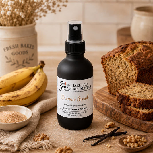 A bottle of Banana Bread Room & Linen Spray | Banana, Walnut & Warm Vanilla by Jarhead Aromatics rests on a table surrounded by sliced banana bread, fresh bananas, sugar, vanilla beans, and walnuts—creating a cozy and inviting kitchen scene that captures the comforting essence of home.