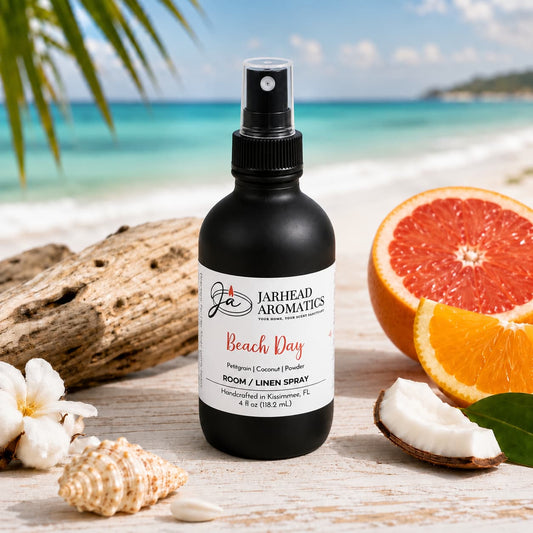 A bottle of Jarhead Aromatics Beach Day Room & Linen Spray | Citrus, Coconut & Coastal Musk rests on the sand among seashells, sliced grapefruit, coconut pieces, and palm leaves, with the ocean and sky softly blurred in the background—evoking the ideal setting for a beach-inspired home fragrance.