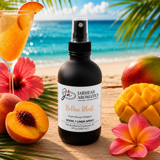 A bottle of Jarhead Aromatics Bellini Blast Room & Linen Spray | Peach, Mango & Passionfruit rests on a beachside table, surrounded by a tropical drink, fresh mango slices, vibrant flowers, and palm leaves. The blue ocean and sky form the perfect backdrop to showcase this tropical home fragrance.