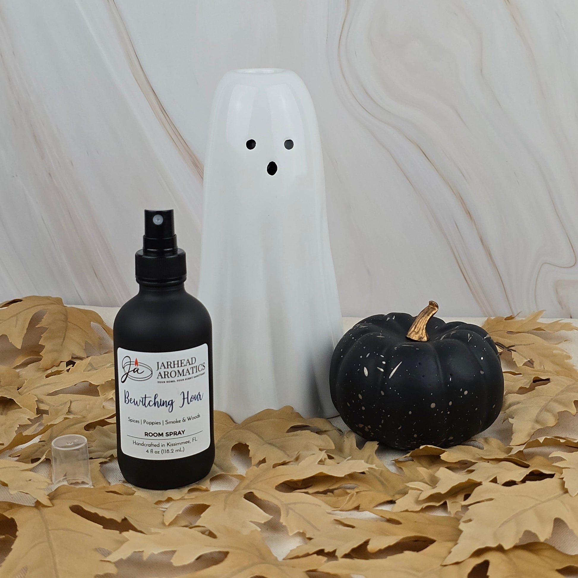 A white ceramic ghost, a black pumpkin with gold specks, and Jarhead Aromatics’ Bewitching Hour Room Spray from the Halloween Collection sit on tan leaves against a marble-patterned background.