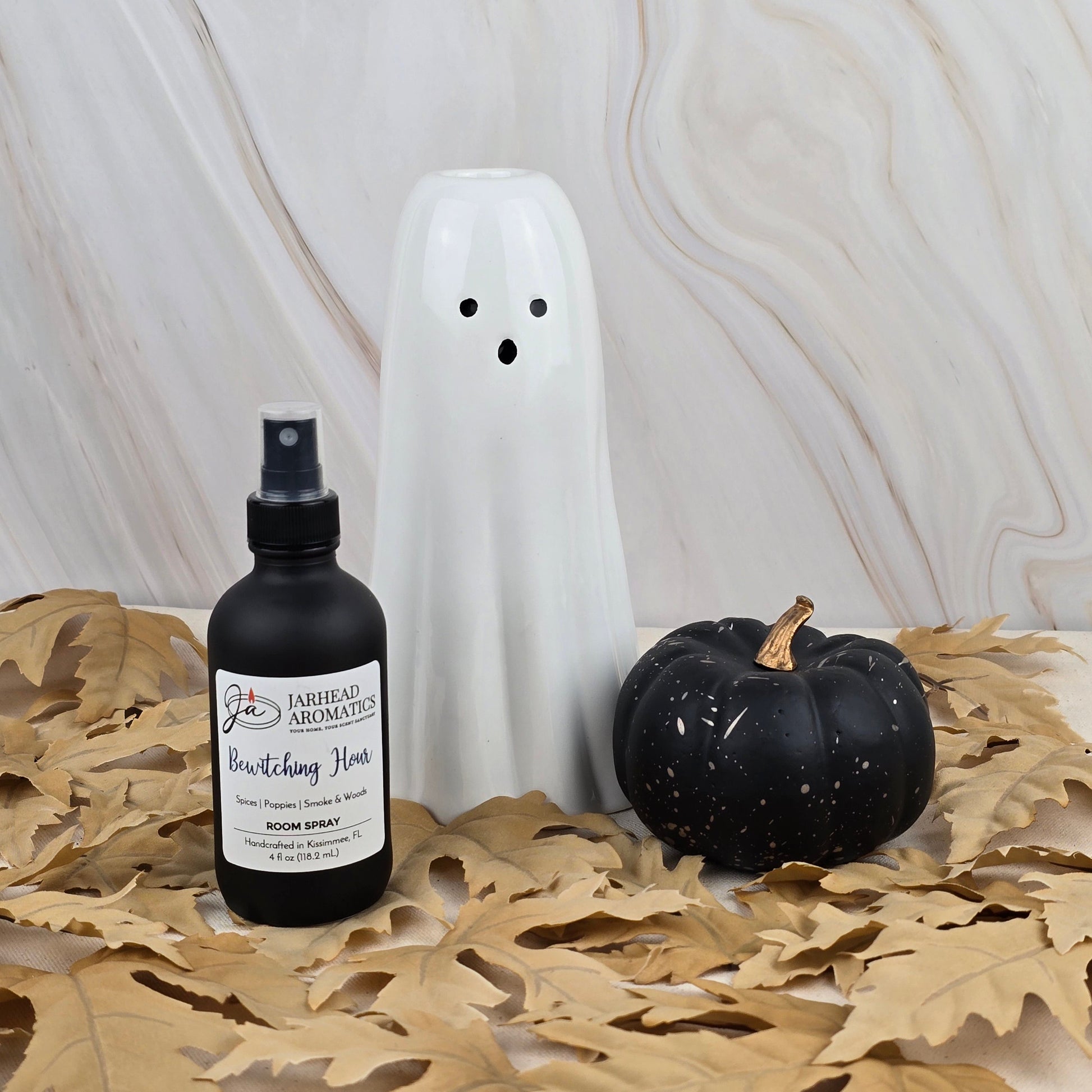 A white ghost figurine, a black pumpkin, and Jarhead Aromatics’ Bewitching Hour - Room Spray from the Halloween Collection with an enchanting floral scent are displayed on dried leaves against a marble background.