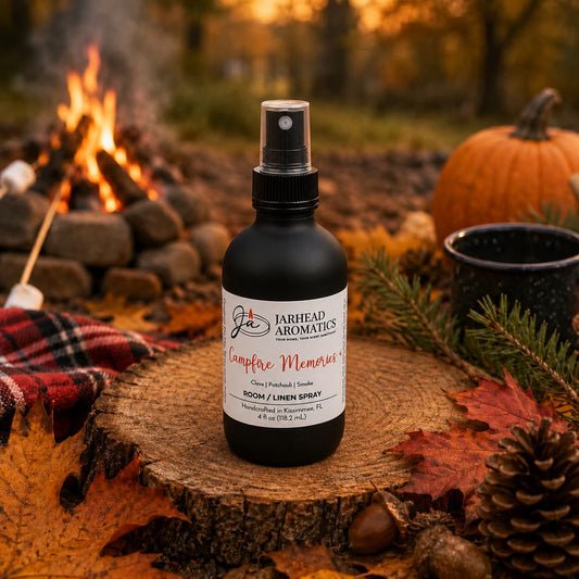 A bottle of Jarhead Aromatics Campfire Memories Room & Linen Spray (Maple, Clove & Smoky Embers) rests on a wooden surface outdoors, surrounded by autumn leaves, a pumpkin, and pinecones, with marshmallows roasting over a campfire in the background—capturing the essence of cozy home fragrance perfect as a campfire-inspired room spray.